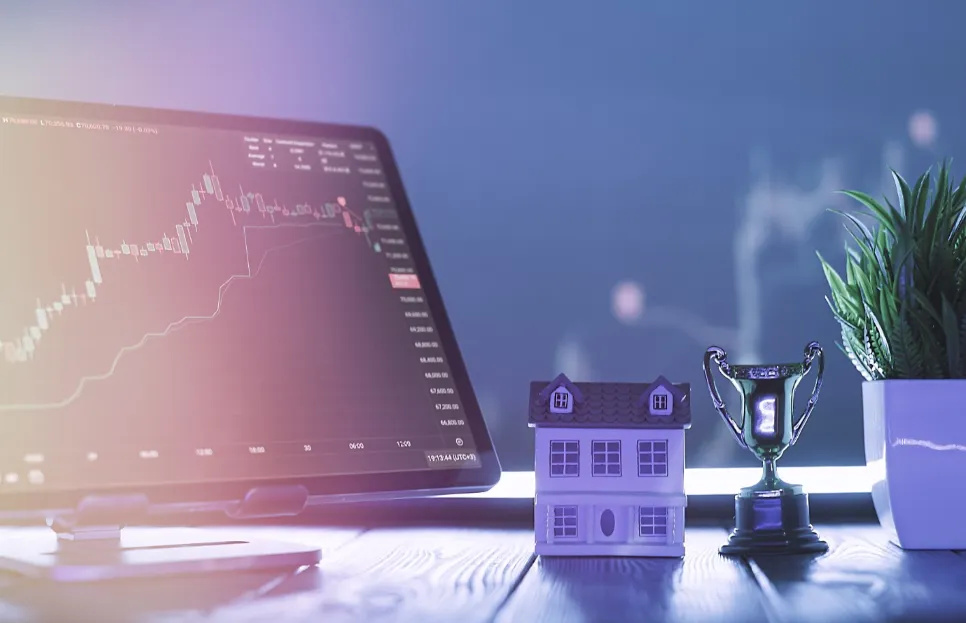 Tablet s grafom rastu na obrazovke, vedľa neho model domu, malá trofej a izbová rastlina na stole