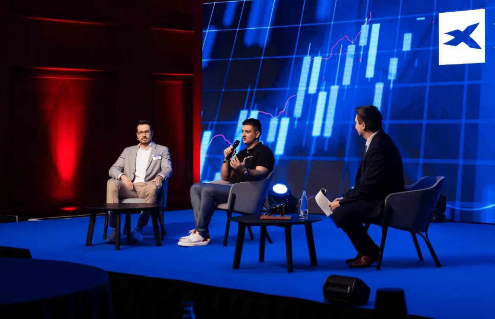 Panelová diskusia na Online Trading konferencii 2026 – traja muži sedia na pódiu v kreslách, jeden hovorí do mikrofónu, za nimi je veľká obrazovka s grafom finančných trhov a červené/modré osvetlenie.