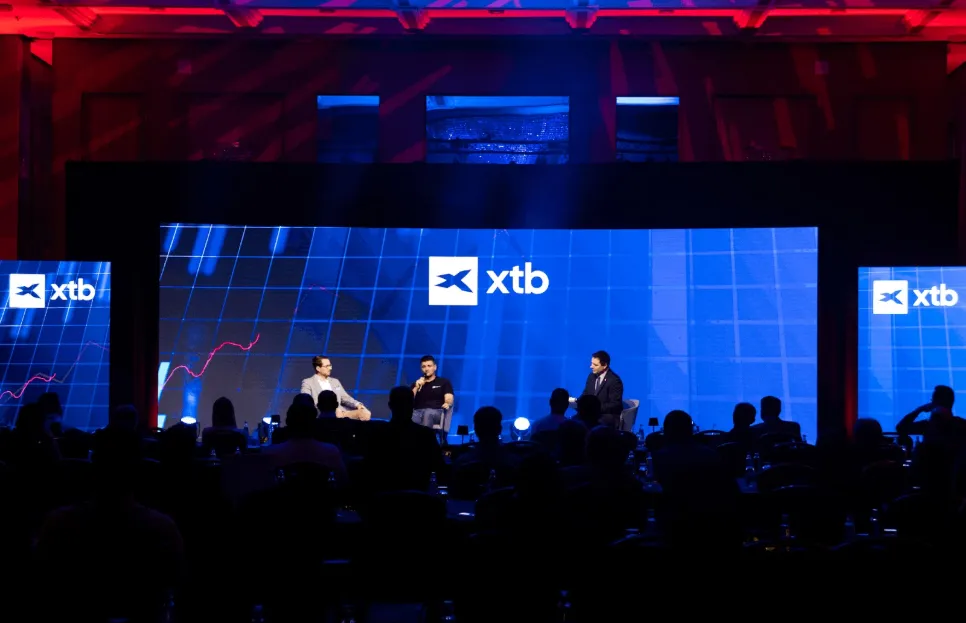 Panelová diskusia na konferencii XTB, traja rečníci sedia na pódiu pred veľkou obrazovkou s logom XTB a grafom, publikum sedí v tmavej miestnosti